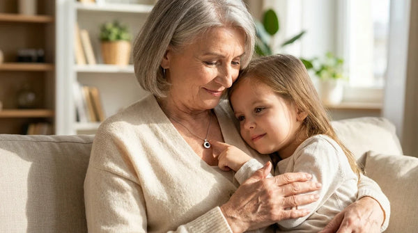 A grandmother in a cream knit cardigan seated with her young granddaughter on her lap, the granddaughter gently touching a small Brother & Sisters Circle Photo Necklace pendant at the grandmother's clavicle